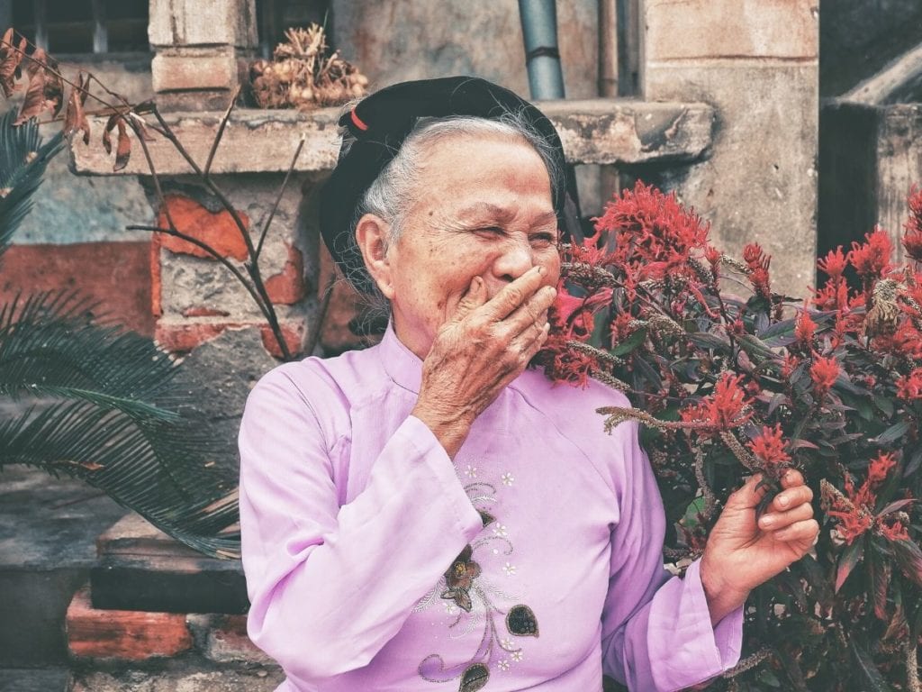 An old woman smiles while holding petaled flowers.