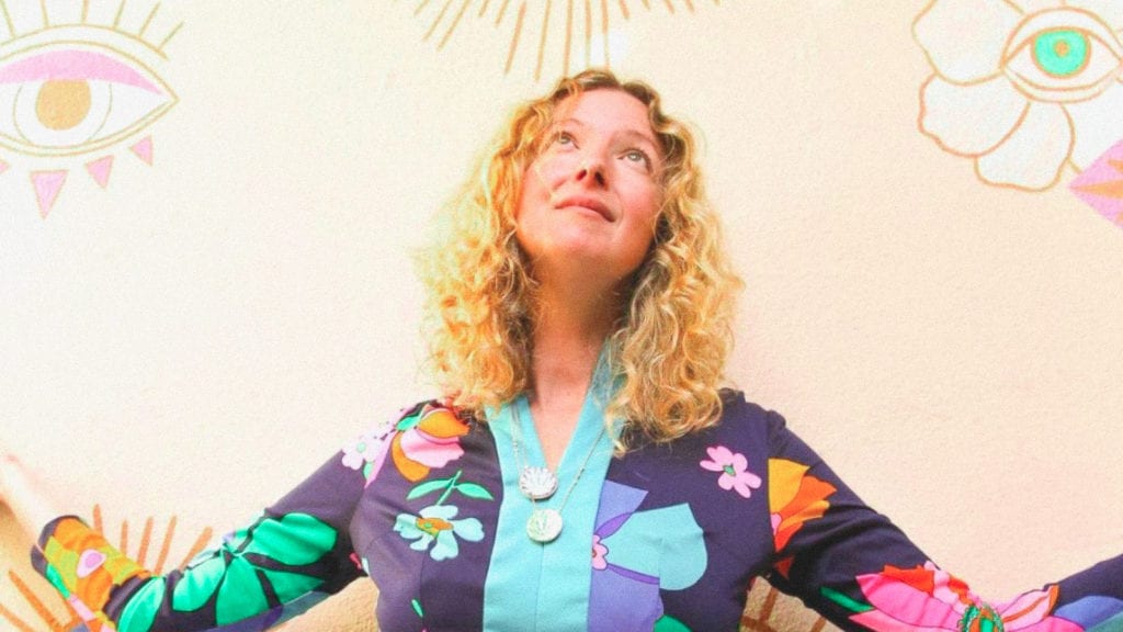 A photograph of Zoe Wilder standing in front of a psychedelic wall wearing psychedelic clothing.