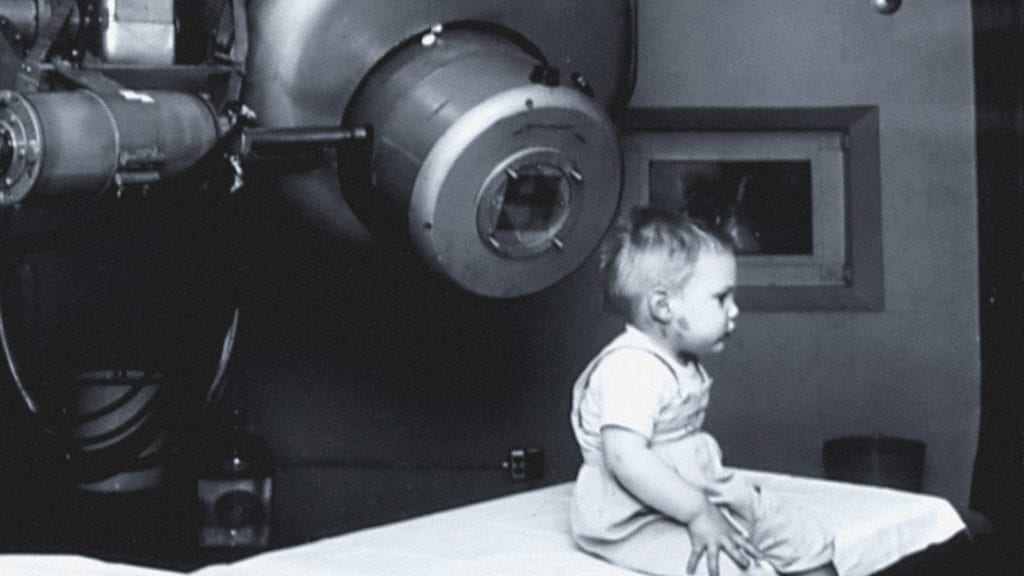 A vintage image of a child sitting on a hospital bed with machinery around him, depicting paediatrics.