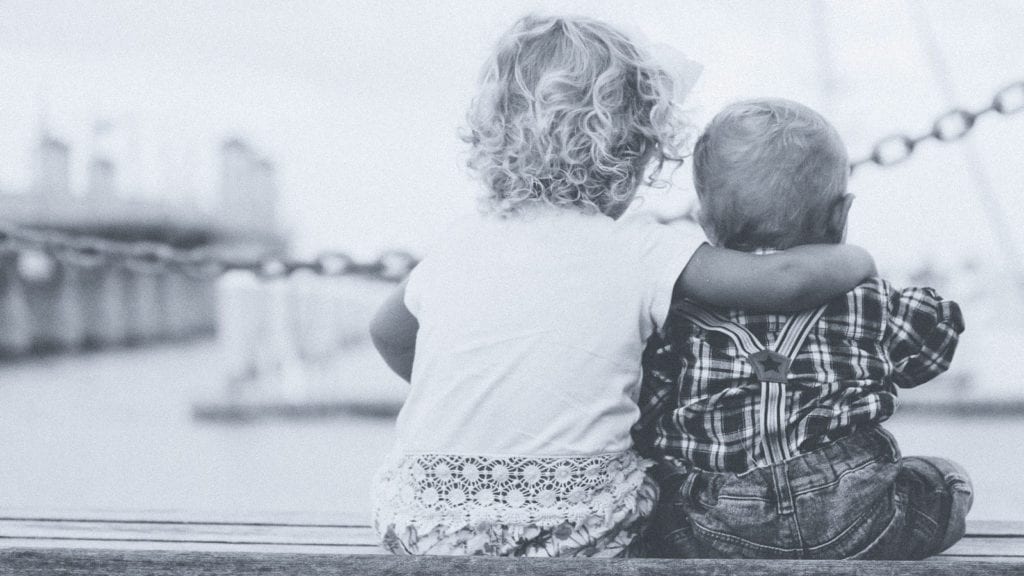 A photograph of two children sitting on a wooden bench, hugging each other. 