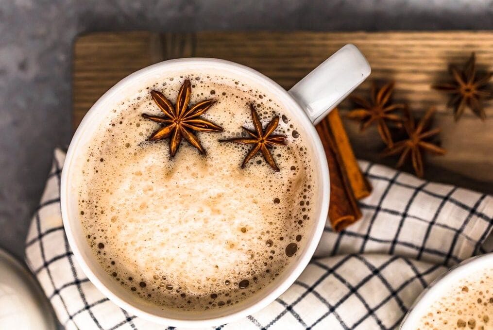 A glass of milky spiced chai surrounded by peppercorns
